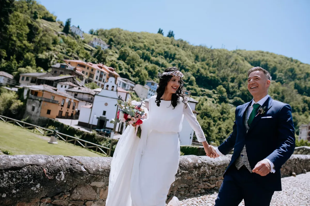 Novios cogidos de la mano sonriendo cruzando el punte romano de Cangas del Narcea